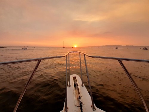 View from the bow of a boat at sunset, with calm waters and silhouettes of distant boats against a colorful sky.