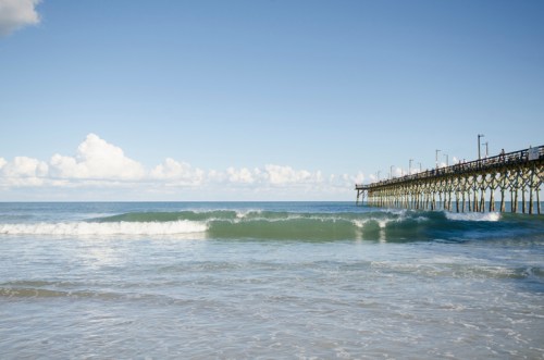 A serene beach scene featuring gentle waves and a wooden pier extending into the calm ocean under a clear blue sky.