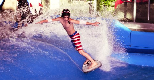 A boy in swim trunks rides a flowboard, creating a splash of water in a vibrant indoor water park.