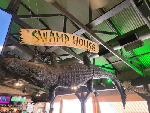 A large alligator sculpture hangs from the ceiling under a sign that reads "Swamp House" in a rustic setting.