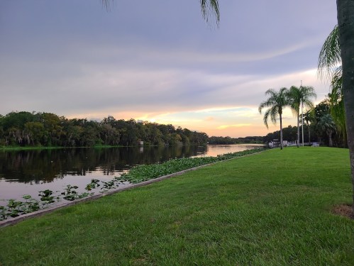 A serene river scene at sunset, with lush greenery and palm trees lining the water's edge.