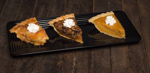 Three slices of pie on a black plate: apple, pecan, and pumpkin, each topped with a dollop of whipped cream.