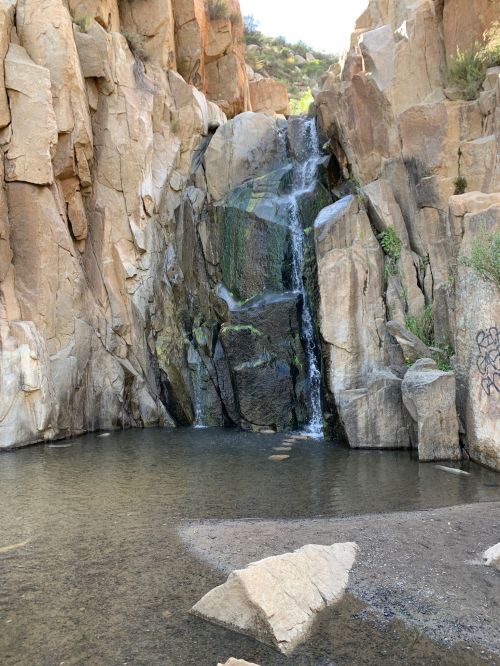 A serene waterfall cascading over rocky cliffs into a clear pool, surrounded by rugged terrain and sparse vegetation.