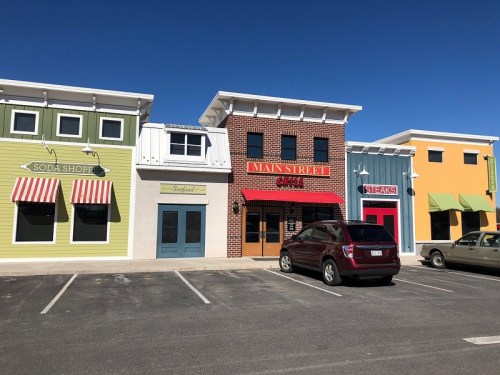 Colorful storefronts line a street, featuring a soda shop, seafood restaurant, and a steakhouse, with parked cars.
