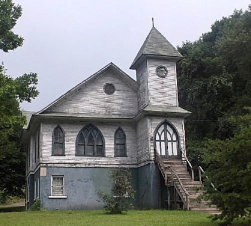 A weathered, two-story church with a steeple, arched windows, and a staircase, surrounded by greenery.