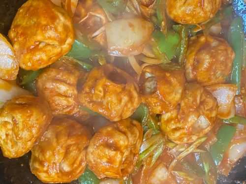 A close-up of meatballs in a spicy sauce with green peppers and onions, steaming in a pan.
