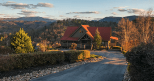 A scenic view of a house with a red roof, surrounded by trees and mountains at sunset.