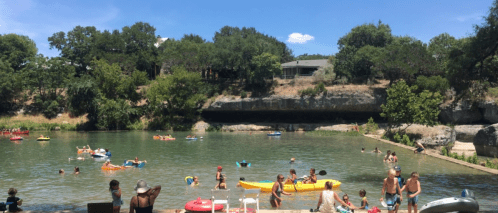 A sunny day at a river with people kayaking, swimming, and relaxing by the shore among trees and rocks.