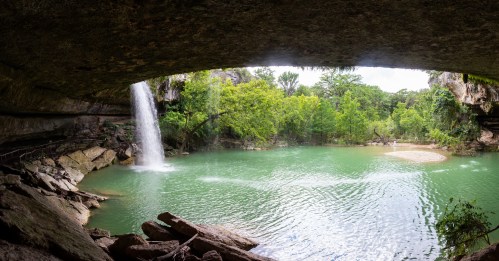 A serene waterfall cascades into a green pool, surrounded by lush trees and rocky formations under a natural arch.
