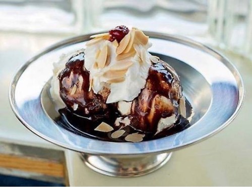 A silver dish holds a scoop of ice cream topped with chocolate sauce, whipped cream, almonds, and a cherry.