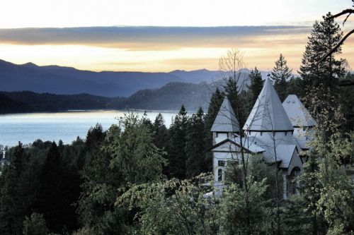 A scenic view of a lake surrounded by mountains, featuring a castle-like structure amidst lush trees at sunset.