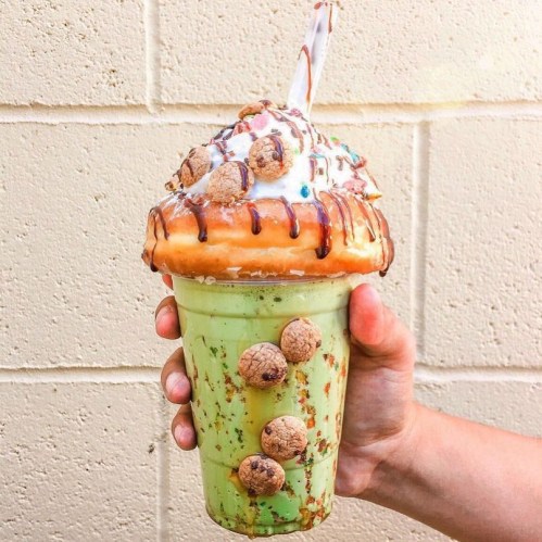 A hand holding a large, colorful milkshake topped with whipped cream, a donut, and cookie pieces.