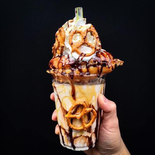 A hand holding a tall dessert cup filled with ice cream, topped with whipped cream, pretzels, and chocolate drizzle.