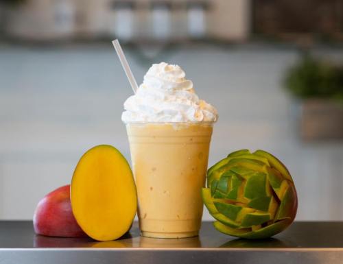 A creamy mango smoothie topped with whipped cream, surrounded by a whole mango and a sliced green mango.