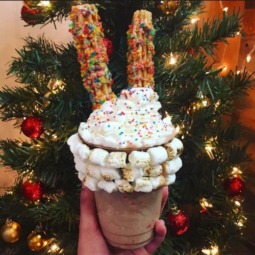 A festive milkshake topped with whipped cream, colorful sprinkles, and two churros, held in front of a Christmas tree.