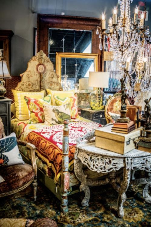 A cozy bedroom setup featuring a colorful bed, decorative pillows, vintage furniture, and an ornate chandelier.