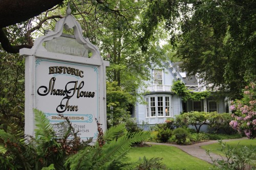 Historic Shan House Inn sign in front of a charming blue house surrounded by lush greenery and blooming flowers.