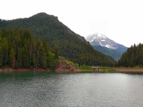 A serene lake surrounded by lush green mountains, with a snow-capped peak in the background under a cloudy sky.