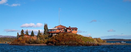 A scenic view of a wooden house on a small island, surrounded by water and trees under a clear blue sky.