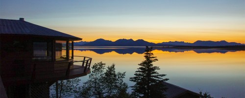Sunset over a calm lake, reflecting mountains and trees, with a cabin in the foreground.