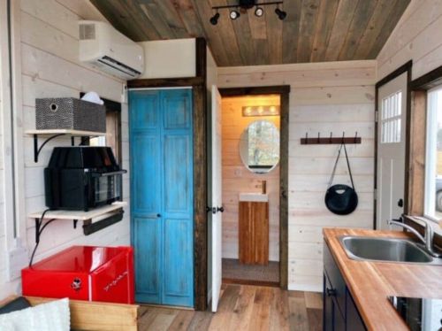 Cozy tiny home interior featuring a blue cabinet, kitchenette, and a bathroom with a circular mirror. Warm wood accents.