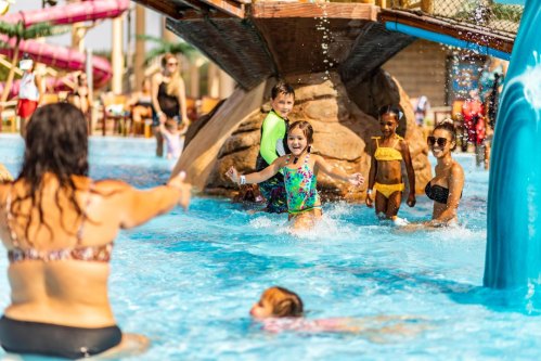 A group of children and adults enjoy a water park, splashing in the pool near a water slide on a sunny day.