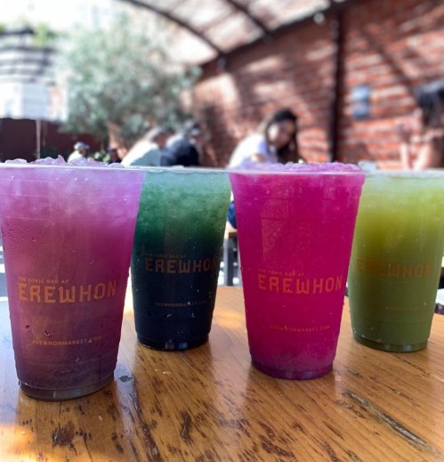 Four colorful drinks in clear cups on a wooden table, with a sunny outdoor setting in the background.