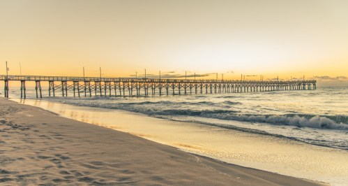 A serene beach scene at sunset featuring a long wooden pier extending over gentle waves.