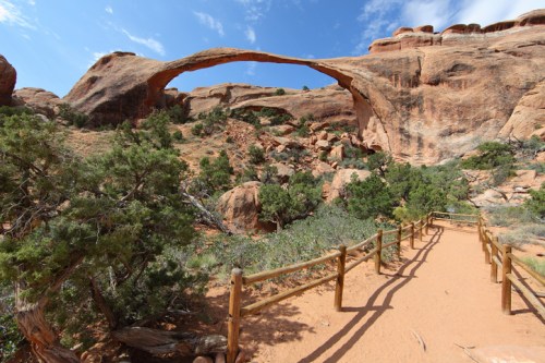 A natural stone arch rises above a sandy path surrounded by greenery and rocky terrain under a blue sky.