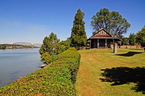 A serene riverside scene featuring a house, lush greenery, and a bridge in the background under a clear blue sky.