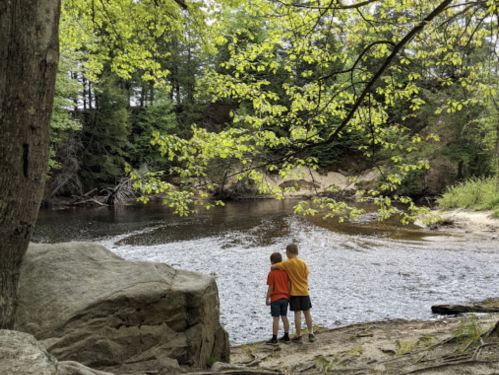 Two children stand by a calm river, surrounded by lush green trees and rocks, enjoying a peaceful moment together.