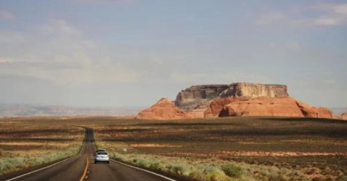 A car drives down a long road with red rock formations and a distant plateau under a blue sky.