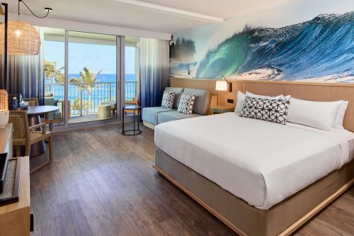 A modern hotel room featuring a king bed, ocean view, and beach-themed decor with a large wave mural.