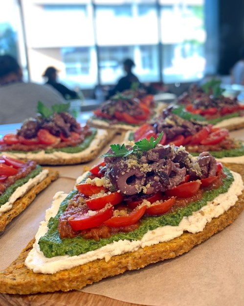 A close-up of open-faced sandwiches topped with pesto, tomatoes, olives, and herbs, served on a wooden table.