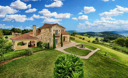 A picturesque stone house on a hillside, surrounded by lush greenery and rolling hills under a blue sky with fluffy clouds.
