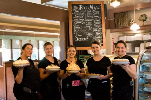 Five women in black shirts hold pies in front of a chalkboard menu in a cozy bakery setting.