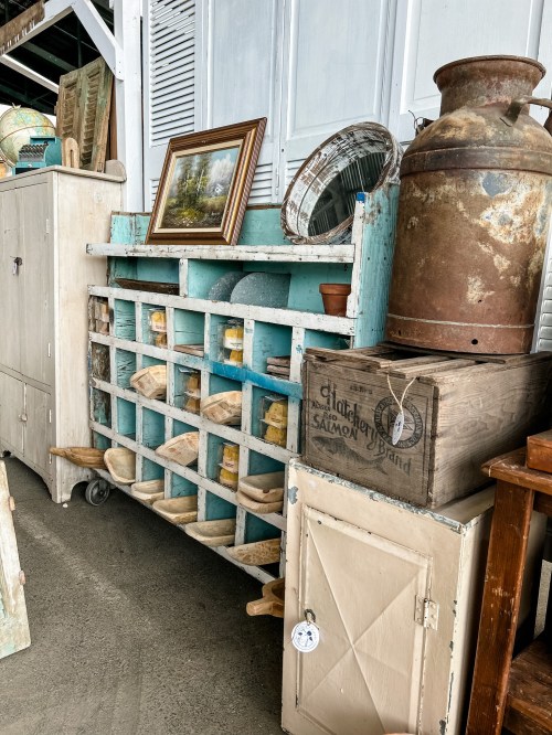 A vintage display with wooden shelves, jars, a mirror, and various rustic items, set in a charming interior space.