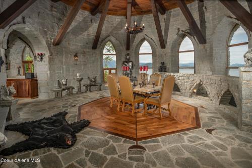 A stone-walled dining area with a wooden table, chairs, and a bear rug, featuring large windows and gothic decor.