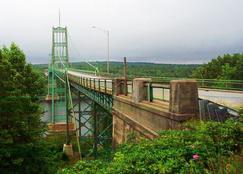 A green suspension bridge spans a river, surrounded by lush greenery and a cloudy sky in the background.