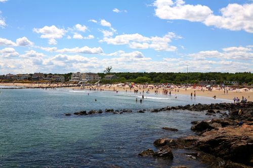 A sunny beach scene with people walking along the shore, rocky outcrops, and clouds in a blue sky.