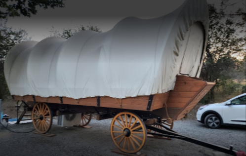 A covered wagon with a wooden frame and large wheels, parked on gravel near a white vehicle.