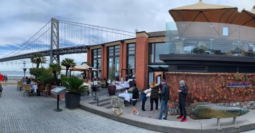 Outdoor dining area with people, palm trees, and a view of a bridge in the background. Cloudy sky above.