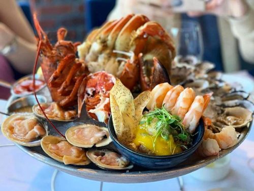 A seafood platter featuring lobster, shrimp, oysters, clams, and a mango garnish, served on a round metal tray.