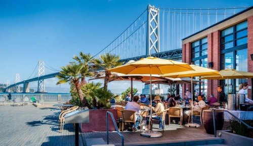 Outdoor dining area with umbrellas, palm trees, and a view of a bridge against a clear blue sky.