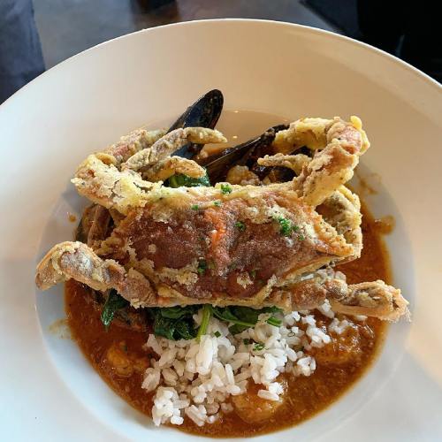 A plate of fried soft-shell crab atop rice, surrounded by mussels and a rich sauce, garnished with herbs.