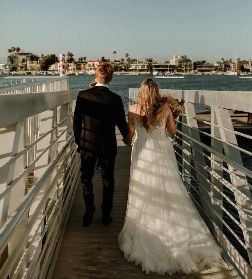 A couple walks hand in hand along a pier, with the ocean and palm trees in the background, dressed in wedding attire.