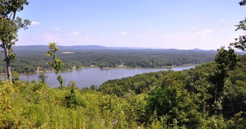 A panoramic view of a serene lake surrounded by lush green forests and distant hills under a clear blue sky.