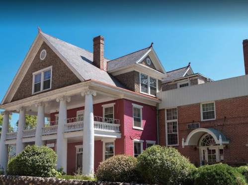 A large, historic house with a pink facade, white columns, and a modern addition, surrounded by greenery under a clear blue sky.