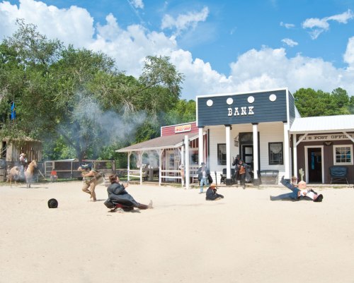 A Wild West scene with cowboys in a shootout outside a bank, dust and smoke in the air, and horses nearby.