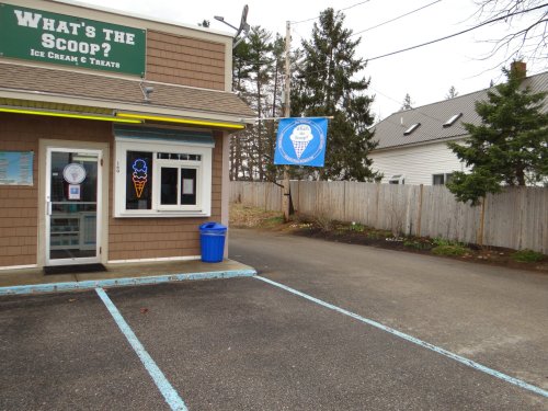 Exterior of an ice cream shop named "What's the Scoop?" with a blue ice cream sign and a parking area.